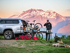 Camping scene of man cooking next to his truck. Bikes and mountains in the background